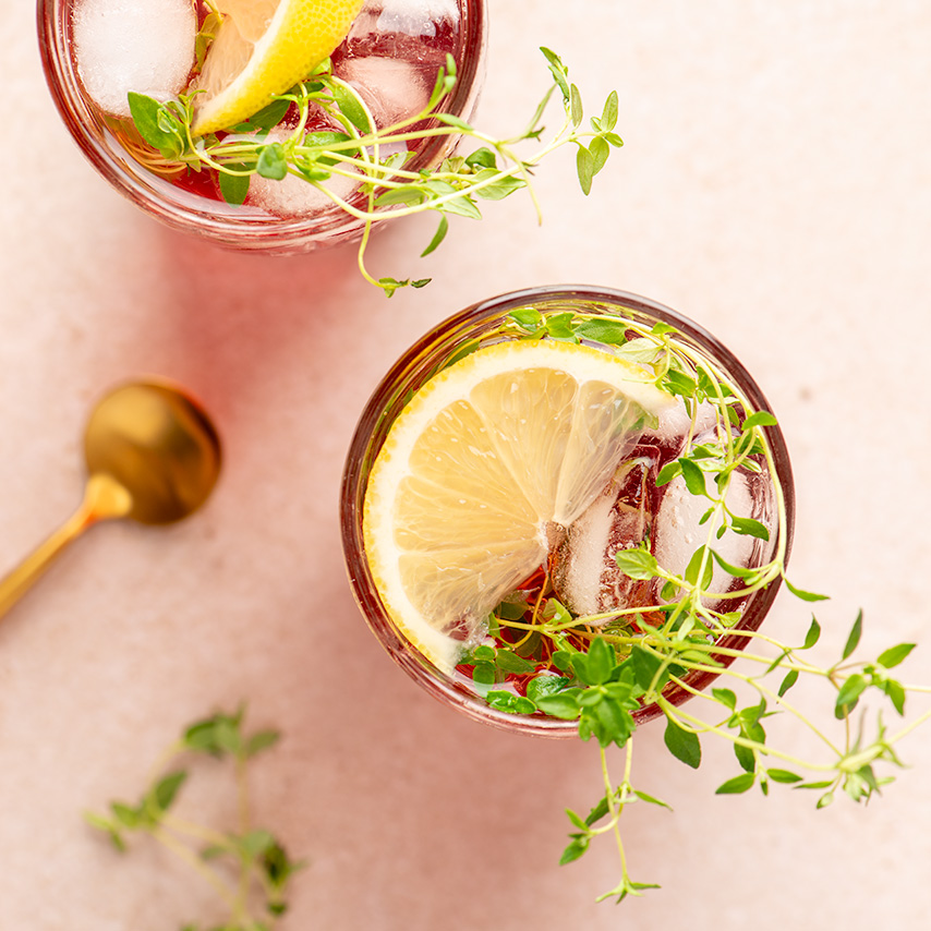 Two red drinks shown from above with sliced lemon and fresh thyme inside, a gold spoon on the counter next to them